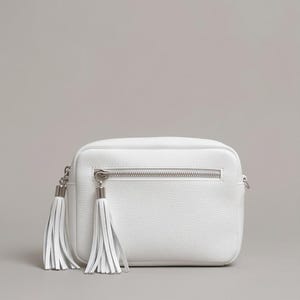 Puede incluir: Un bolso bandolera rectangular de color blanco con textura granulada, que cuenta con un bolsillo frontal horizontal con cremallera y dos largas borlas de cuero blanco unidas a los tiradores plateados. Dise&ntilde;o estructurado con herrajes plateados.