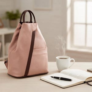 May include: A blush pink backpack with brown accents, a front pocket, and a drawstring closure. The bag has brown handles and a brown vertical stripe. A white mug of coffee, a notebook, and a pen are on the table.