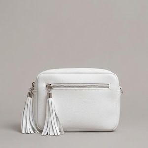 Puede incluir: Un bolso bandolera rectangular de color blanco con textura granulada, que cuenta con un bolsillo frontal horizontal con cremallera y dos largas borlas de cuero blanco unidas a los tiradores plateados. Dise&ntilde;o estructurado con herrajes plateados.