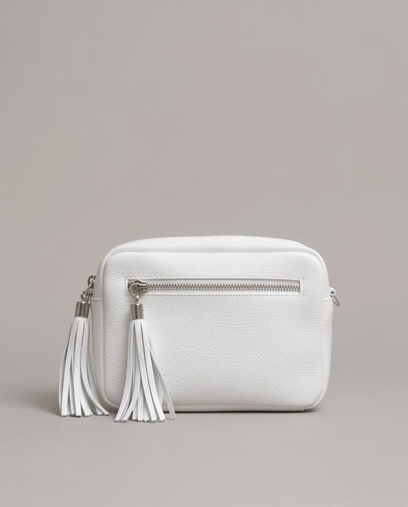 Puede incluir: Un bolso bandolera rectangular de color blanco con textura granulada, que cuenta con un bolsillo frontal horizontal con cremallera y dos largas borlas de cuero blanco unidas a los tiradores plateados. Dise&ntilde;o estructurado con herrajes plateados.