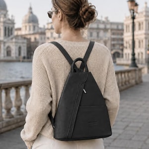 May include: A black triangular backpack with a top handle, adjustable shoulder straps, and a vertical zippered front pocket. The bag features a smooth matte finish and a small embossed logo on the lower front section, providing a structured design.