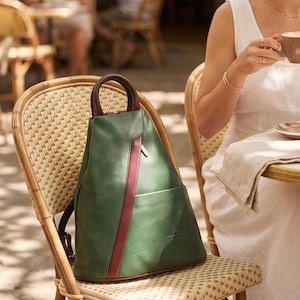 May include: A forest green backpack with a smooth finish, featuring a dark burgundy vertical stripe down the center and a matching dark burgundy rounded top handle. The bag has a structured, triangular silhouette with a front slip pocket.
