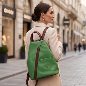 May include: A bright green leather backpack featuring dark brown leather trim, a top handle, and adjustable shoulder straps. The bag has a structured, tapered silhouette with a front slip pocket and visible stitching along the brown leather accents.