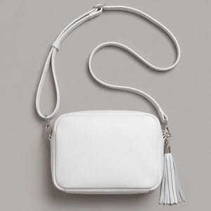 Puede incluir: Un bolso bandolera rectangular de color blanco con superficie texturizada, que cuenta con una correa larga ajustable y un herraje de metal plateado en el lateral que sostiene una borla de cuero blanco a juego con el dise&ntilde;o del bolso.