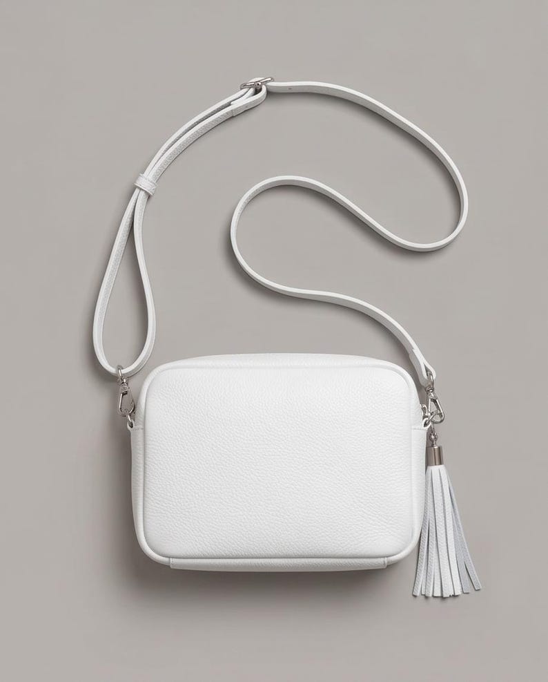 Puede incluir: Un bolso bandolera rectangular de color blanco con superficie texturizada, que cuenta con una correa larga ajustable y un herraje de metal plateado en el lateral que sostiene una borla de cuero blanco a juego con el dise&ntilde;o del bolso.