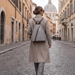 May include: A light grey backpack with dark brown leather straps and a diagonal dark brown leather accent across the front. The backpack features a top handle, a structured triangular shape, and a smooth finish, worn over a beige wool coat and grey trousers.