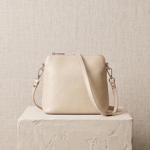 May include: A smooth, cream-coloured crossbody bag with a rounded top silhouette, a top zip closure, and a matching adjustable shoulder strap attached with silver-toned metal hardware clips on both sides of the structured bag body.