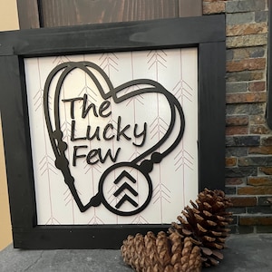 May include: Black wooden framed sign with a white background and a black heart shape with the text "The Lucky Few" inside. The heart shape has a black circle with three black arrows pointing upwards inside. The background has a pattern of small pink arrows.