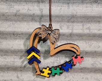 Può includere: Ciondolo a forma di cuore in legno con un fiocco marrone e pezzi di puzzle in blu, verde, giallo e rosso. I pezzi del puzzle sono disposti in un cerchio attorno al cuore. Il ciondolo è appeso a uno spago marrone.