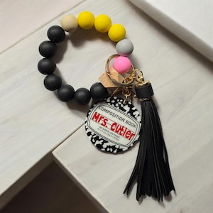 May include: Black silicone bracelet with yellow, beige, and gray beads. Attached to the bracelet is a keychain with a black tassel, a gold key ring, and a round tag with a black and white composition book design. The tag says "COMPOSITION BOOK" and "Mrs. Cutler" in red text. The tag also says "100 SHEETS-200 PAGES" and "9 7/7 in/24.7-19.5cm".