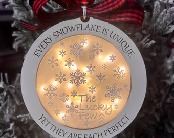 May include: White wooden Christmas ornament with a gold snowflake design and the text "Every snowflake is unique, the lucky few, yet they are each perfect". The ornament has a red and white checkered ribbon and a small red and white checkered ball at the top.