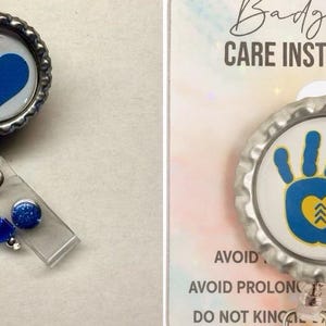 May include: A silver badge reel with a blue heart design and a DNA charm. The reel has a clear plastic ID holder and a blue glitter accent. The badge reel is a functional accessory.