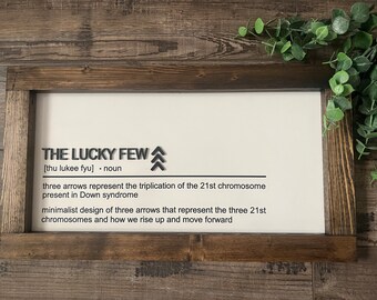 May include: A white framed sign with the text "THE LUCKY FEW [thu lukee fyu] . noun" and a black illustration of three arrows pointing upwards. The text below the title reads "three arrows represent the triplication of the 21st chromosome present in Down syndrome minimalist design of three arrows that represent the three 21st chromosomes and how we rise up and move forward"