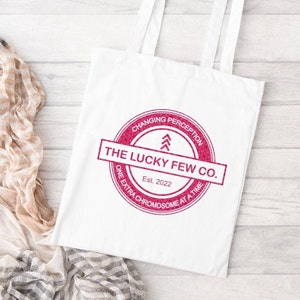 May include: White tote bag with a pink circle logo that says "CHANGING PERCEPTION" and "THE LUCKY FEW CO." with the text "Est. 2022" and "ONE EXTRA CHROMOSOME AT A TIME" underneath.