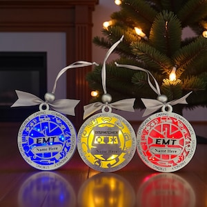 May include: Three illuminated Christmas ornaments with silver ribbons. One is blue with "EMT" and "Name Here", another is yellow with "Dispatcher" and "Name Here", and the third is red with "EMT" and "Name Here". Each has words like "Caring" and "Dependable".