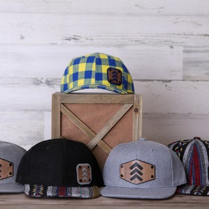 May include: Five baseball caps with different patterns and colors. Each cap has a wooden patch with an arrow design. The caps are arranged on a wooden surface in front of a white wood plank wall.