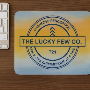 May include: A mousepad with a watercolor design in yellow, blue, and orange. The mousepad features the text "THE LUCKY FEW CO." in a rectangular box, and a circular design with the text "CHANGING PERCEPTION" and "ONE EXTRA CHROMOSOME AT A TIME". A white mouse and keyboard are also visible.