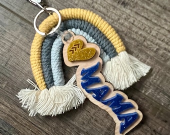 Può includere: Un portachiavi in macramè arcobaleno con un'etichetta in legno che dice "MAMA" in glitter blu. L'etichetta ha anche un piccolo cuore con glitter dorato.