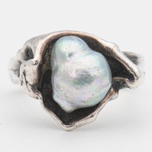 May include: A sterling silver ring featuring a large, irregularly shaped, iridescent pearl. The pearl has a soft, pearlescent sheen with hints of blue and green. The ring's setting has a textured, organic design.