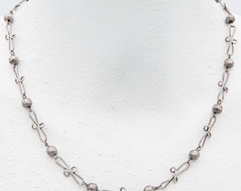 Puede incluir: Collar plateado con un diseño delicado. El collar presenta cuentas de metal martillado intercaladas con eslabones decorativos. El collar se exhibe en un maniquí blanco, mostrando sus intrincados detalles y artesanía.