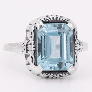 May include: A silver ring with a large, light blue, emerald-cut gemstone set in a filigree design.