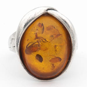 Puede incluir: Anillo de plata con una piedra de ámbar ovalada. El ámbar tiene un color marrón dorado cálido con inclusiones visibles. La banda del anillo tiene un diseño simple y texturizado.