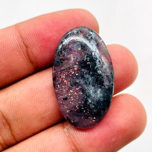 May include: An oval-shaped cabochon gemstone featuring a polished surface with a deep purple and black base colour. The stone displays scattered metallic inclusions that reflect light with iridescent gold, pink, and silver sparkles throughout.