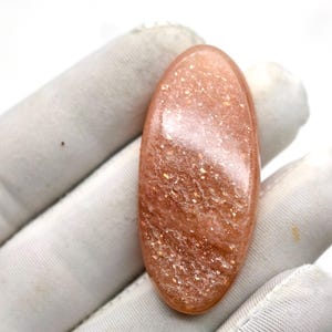 May include: A polished, oval-shaped sunstone gemstone. The stone has a warm, peach-orange hue with shimmering, glitter-like inclusions. The gemstone is held in a white gloved hand, showcasing its natural beauty and unique sparkle.