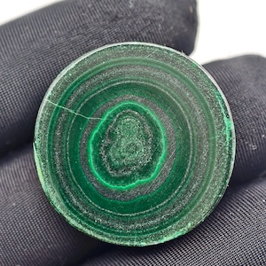 May include: A circular malachite stone, polished to a high sheen, displaying a striking pattern of concentric green bands. The stone's surface features a natural, swirling design with a small crack. The stone is set against a dark backdrop.