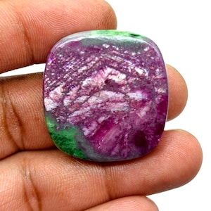 Pode incluir: Uma pedra preciosa polida com formato retangular arredondado. A pedra apresenta uma mistura de cores, incluindo magenta profundo, branco e verde. A área magenta tem uma aparência texturizada e cristalina. A pedra preciosa é segurada em uma mão.