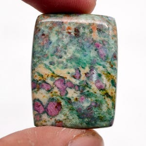 Pode incluir: Uma pedra preciosa retangular com uma mistura de cores. A pedra apresenta uma base de tons verdes e creme, com vibrantes inclusões rosa e vermelhas. A pedra é segurada entre dois dedos.
