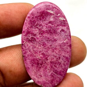 Puede incluir: Una gema ovalada pulida en tonos rosa y magenta. La piedra tiene una superficie texturizada con inclusiones más claras, de color blanco. La gema se sostiene entre los dedos.
