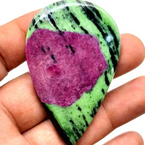 May include: A polished, teardrop-shaped gemstone with a central maroon section and green and black veining. The stone is held in a hand, highlighting its natural colours and patterns. This gemstone is likely used for jewellery or decorative items.