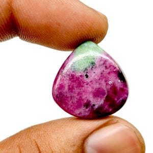 May include: A teardrop-shaped gemstone with a vibrant mix of colours. The stone features a deep magenta base, accented with patches of light green and darker magenta hues. The gem is held between two fingers against a white background.