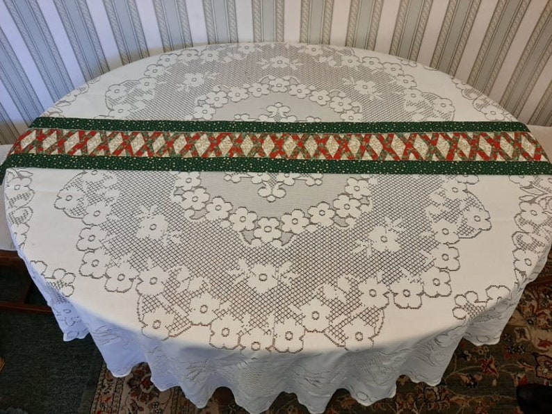 May include: A white lace tablecloth with a green and red patterned table runner. The tablecloth has a floral design.