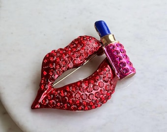 May include: A brooch shaped like a pair of red lips covered in small red crystals. A small lipstick tube with a blue cap and a pink crystal-encrusted base is positioned on the upper right side of the lips. The piece features a gold-tone metal pin.