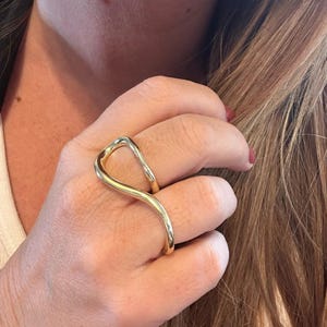 May include: A gold-toned ring with a unique, abstract design. The ring is worn on the index and middle fingers.