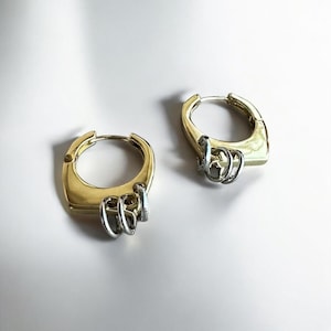 May include: A pair of gold-coloured hoop earrings with a square shape and silver-coloured dangling accents. The earrings are displayed on a white surface, highlighting their design and metallic finish.