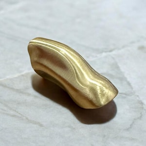 May include: A smooth, curved, gold-coloured metal ring. The ring is made of a single piece of metal and has a unique, organic shape.