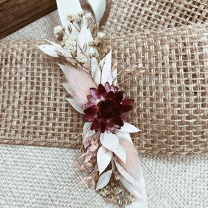 May include: A small bouquet of dried flowers with a burgundy flower, white and pink flowers, and a white ribbon.