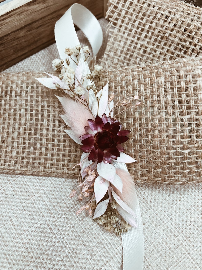 May include: A small bouquet of dried flowers with a burgundy flower, white and pink flowers, and a white ribbon.