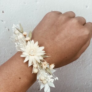 May include: A white dried flower wrist corsage with a large white flower in the center and smaller white and cream flowers surrounding it.