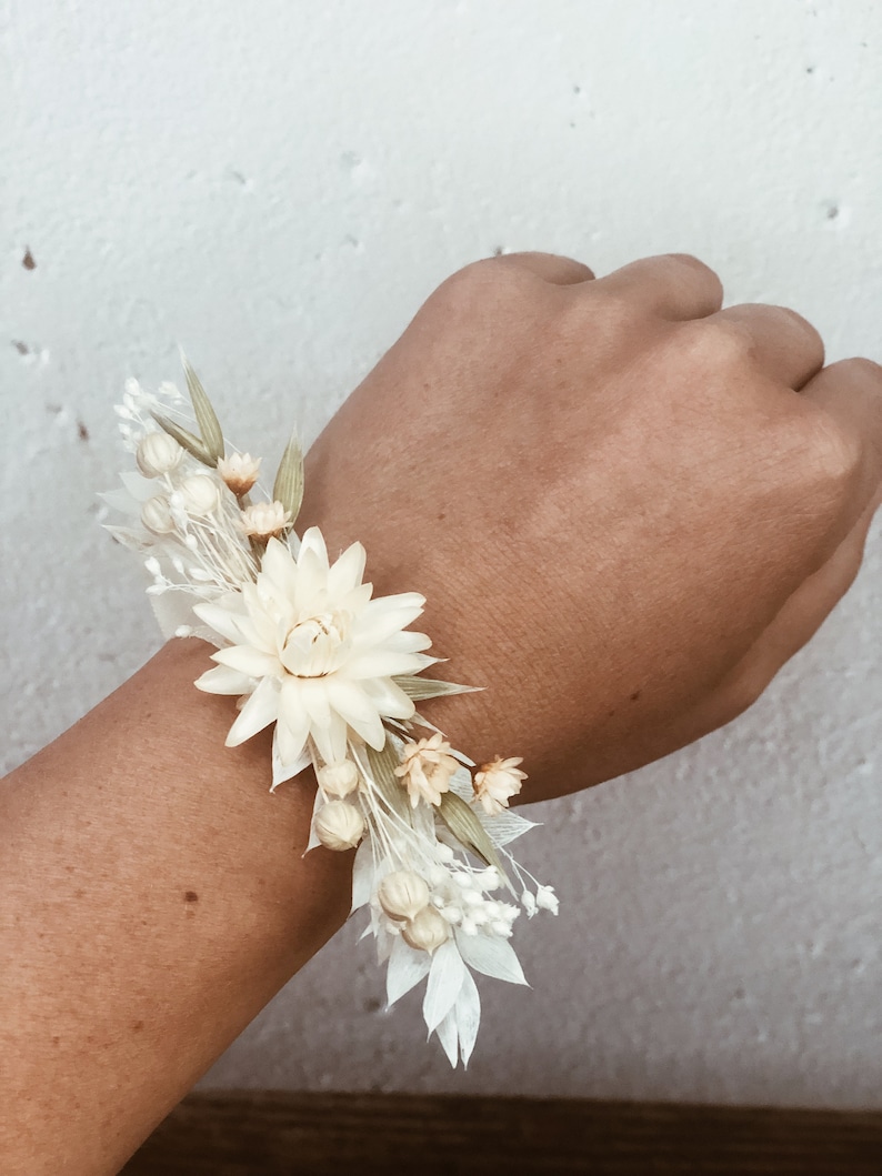 May include: A white dried flower wrist corsage with a large white flower in the center and smaller white and cream flowers surrounding it.