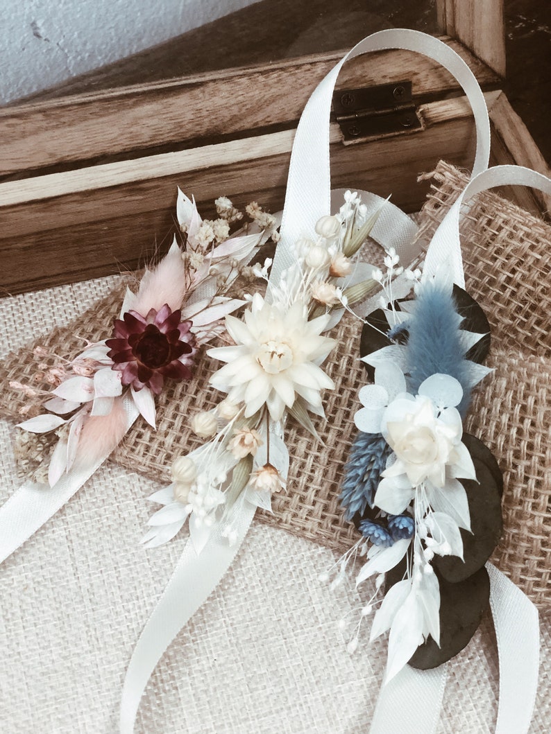 May include: Three dried flower wrist corsages with white satin ribbon. The corsages feature different colors and styles of dried flowers, including white, pink, blue, and burgundy.
