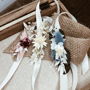 May include: Three white satin ribbon wrist corsages with dried flowers. The corsages feature a variety of dried flowers, including white, cream, pink, burgundy, and blue flowers. The corsages are arranged on a burlap surface.