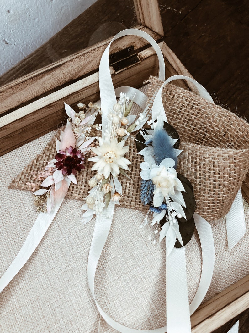 May include: Three white satin ribbon wrist corsages with dried flowers. The corsages feature a variety of dried flowers, including white, cream, pink, burgundy, and blue flowers. The corsages are arranged on a burlap surface.