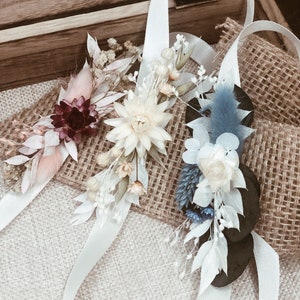 May include: Three white satin ribbons with dried flower arrangements. The arrangements feature white, cream, pink, blue, and burgundy flowers.