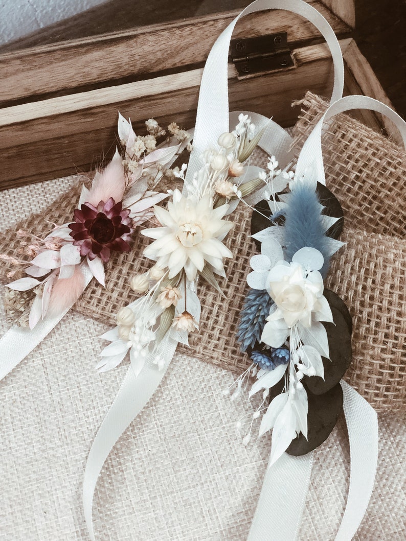May include: Three white satin ribbons with dried flower arrangements. The arrangements feature white, cream, pink, blue, and burgundy flowers.
