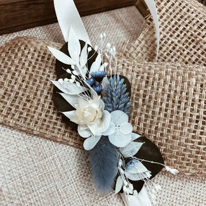 May include: A white ribbon tied around a bouquet of dried flowers. The bouquet includes white, blue, and cream flowers. The bouquet is sitting on a burlap surface.