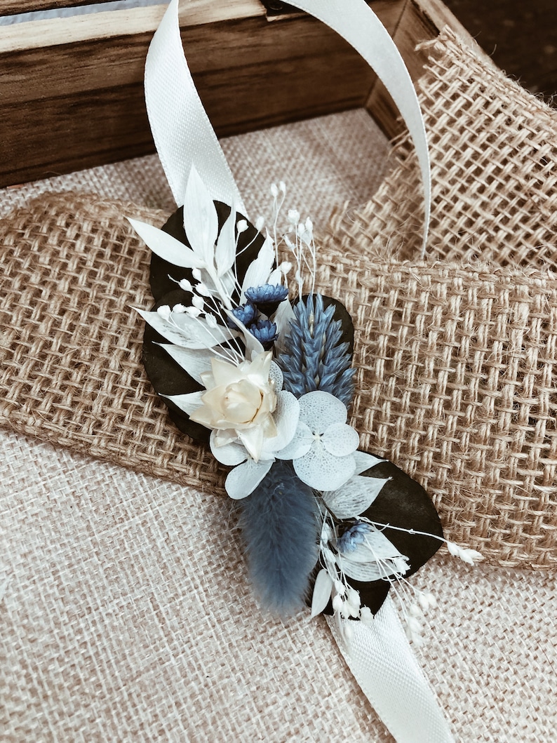 May include: A white ribbon tied around a bouquet of dried flowers. The bouquet includes white, blue, and cream flowers. The bouquet is sitting on a burlap surface.